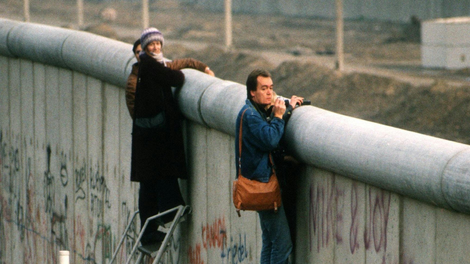 Zwei Personen stehen auf Leitern und gucken über die Berliner Mauer.