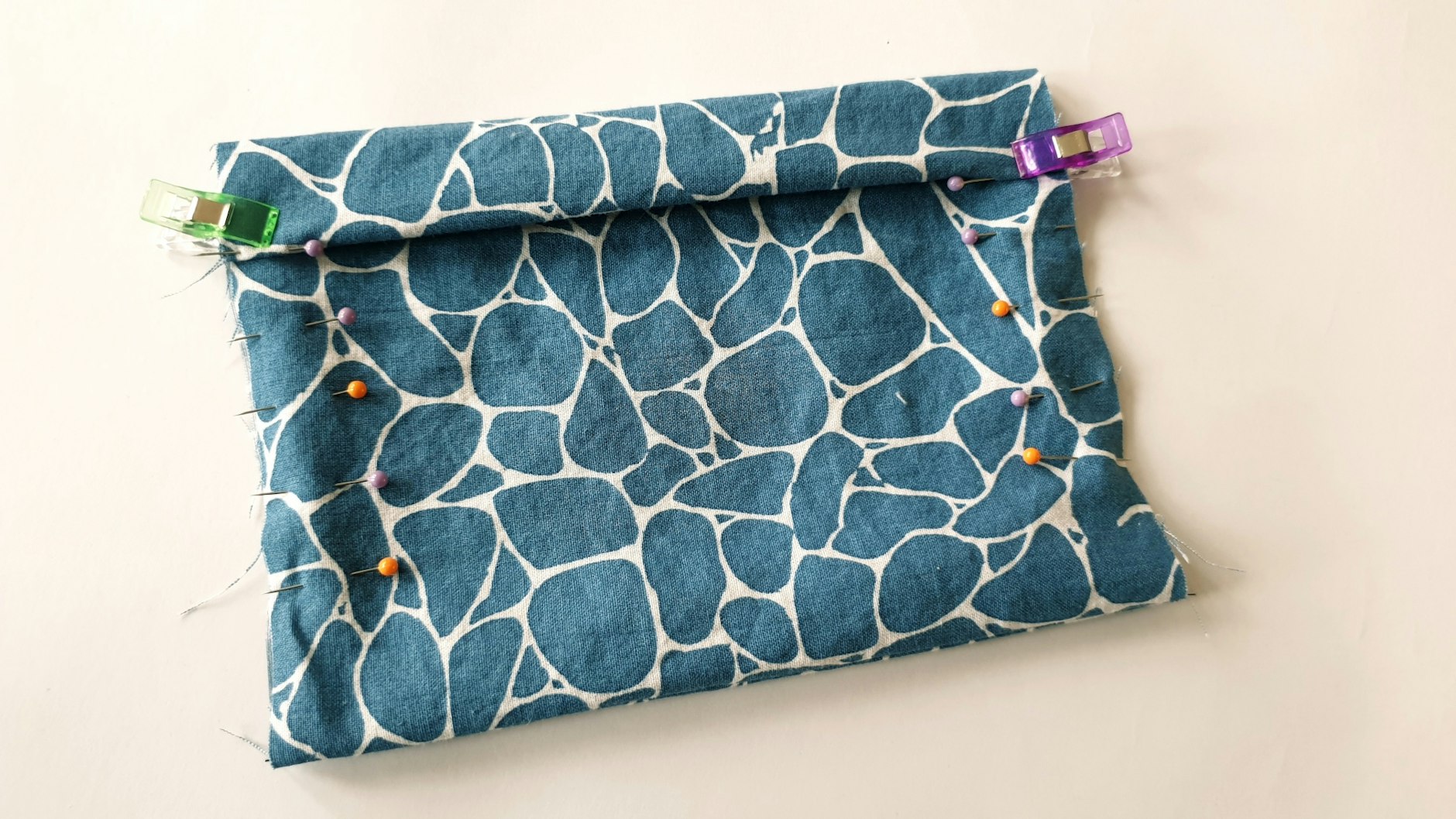 Auf Höhe der ersten Nadeln von oben (hier lilafarben) klappt man den Stoff dann wieder nach oben. Mit zwei Klammern (man kann auch hierfür Stecknadeln verwenden) wird die Falte behelfsmäßig fixiert.