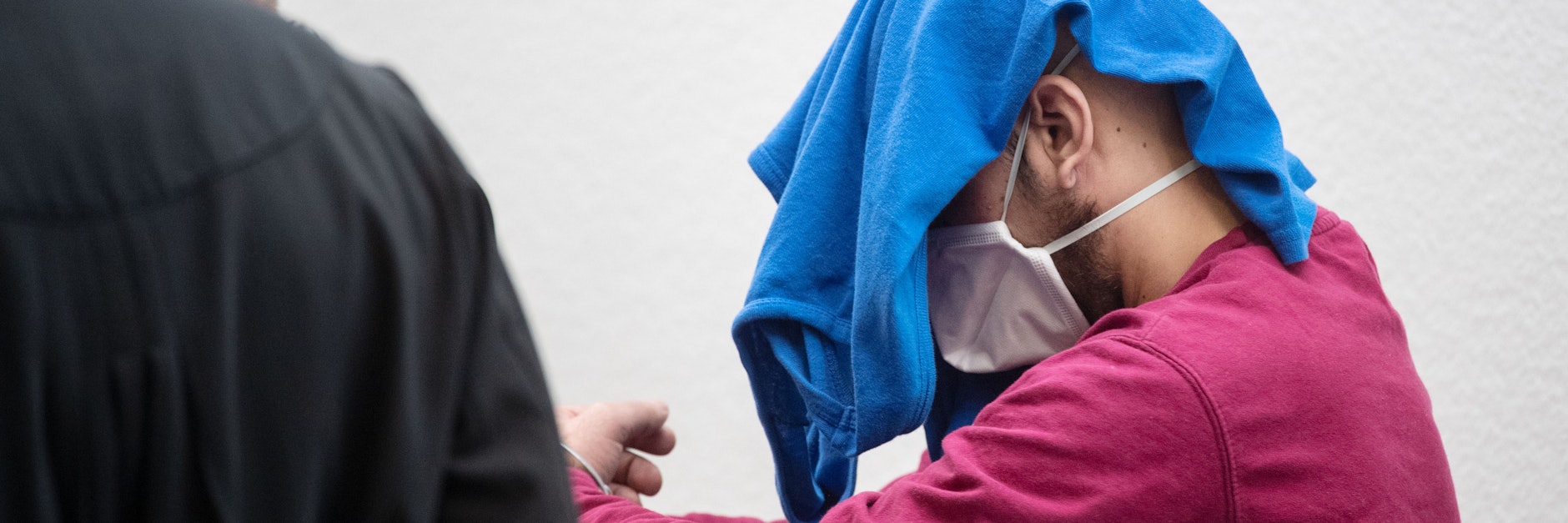 Der Angeklagte trägt eine Schutzmaske als er ins Gericht geführt wird.