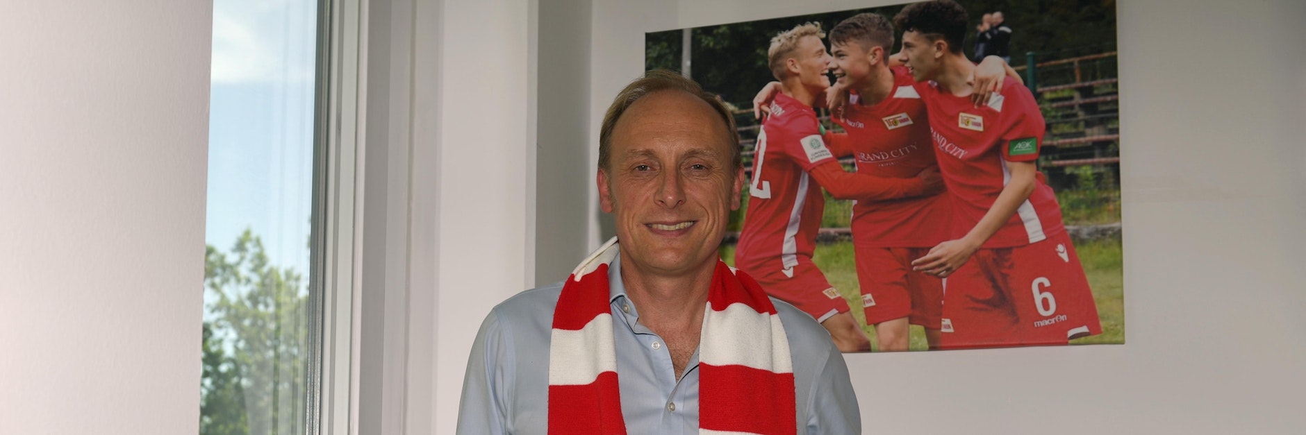 Andrew Wallis in seinem Büro. Im Hintergrund ein Bild von Unions A-Junioren. Dort war er schon engagiert, ehe Aroundtown nach dem Aufstieg bei den Eisernen als Haupt- und Brustsponsor einstieg.&nbsp;