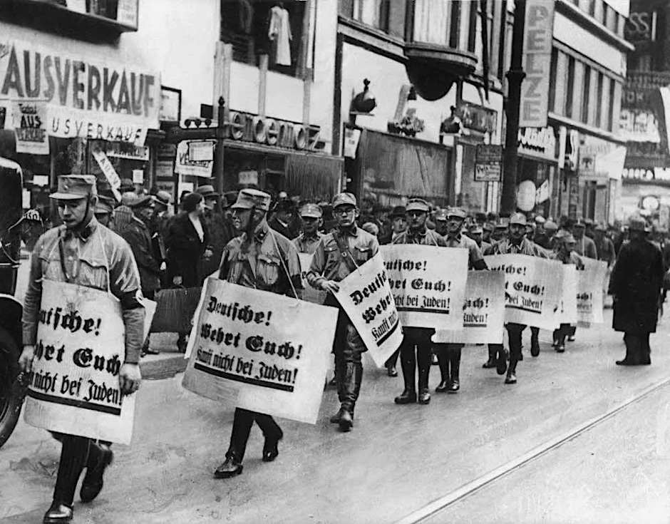 SA-Männer marschieren im November 1938 durch die Straßen von Berlin. Die Plakate tragen die Aufschrift „Deutsche! Wehrt Euch! Kauft nicht bei Juden!“