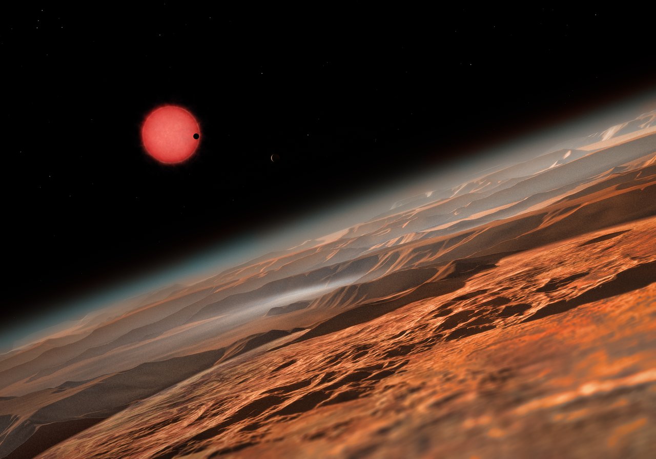 Der rote Zwergstern Trappist-1 ist 40 Lichtjahre von der Erde entfernt. Ihn umkreisen drei Planeten, jeder etwa so groß wie die Erde. Sie befinden sich recht nahe an ihrem Stern, aber weil dieser  deutlich schwächer strahlt als die Sonne, könnte es auf den Planeten bewohnbare Regionen geben. Der Künstler stellt sich vor, wie es auf der Oberfläche eines dieser Welten aussehen mag.