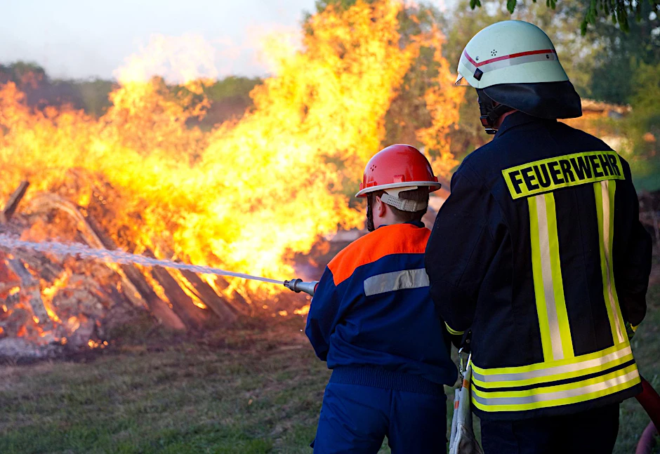 Dieses Bild wird immer seltener: Ein Mitglied der freiwilligen Feuerwehr und ein junges Nachwuchsmitglied halten eine Löschspritze auf ein großes Lagerfeuer im brandenburgischen Sieversdorf.