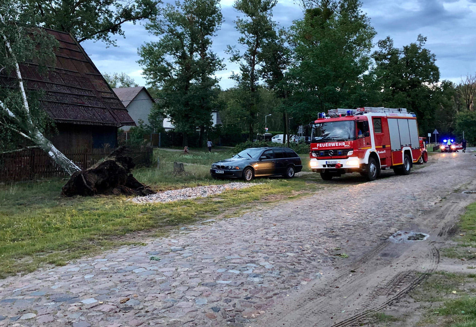 Ein Feuerwehrfahrzeug steht neben einem entwurzelten Baum, die eine Sturmböe auf das Dach eines Hauses gedrückt hat.