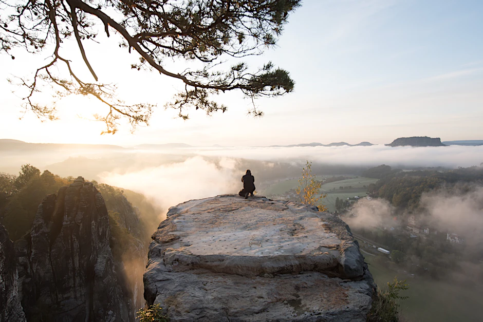 Ein Mann hockt im Nationalpark Sächsische Schweiz auf dem Basteifelsen und fotografiert den Sonnenaufgang.