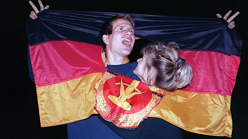 3. Oktober 1990: Ein jubelnden Paar hat sich am Tag der Deutschen Einheit in einer DDR-Fahne dort Platz geschaffen, wo früher das Emblem Hammer und Sichel war