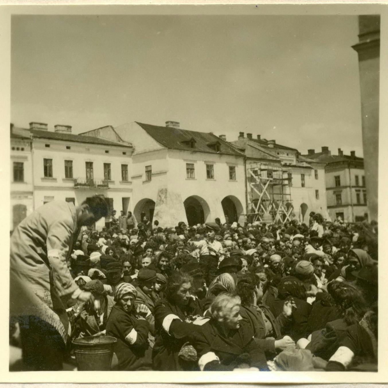 Die Festgehaltenen leiden Durst. Ein Mann mit Armbinde, der möglicherweise zum jüdischen Ordnungsdienst gehört, schöpft Wasser mit einem Becher aus einem herbeigebrachten Eimer.