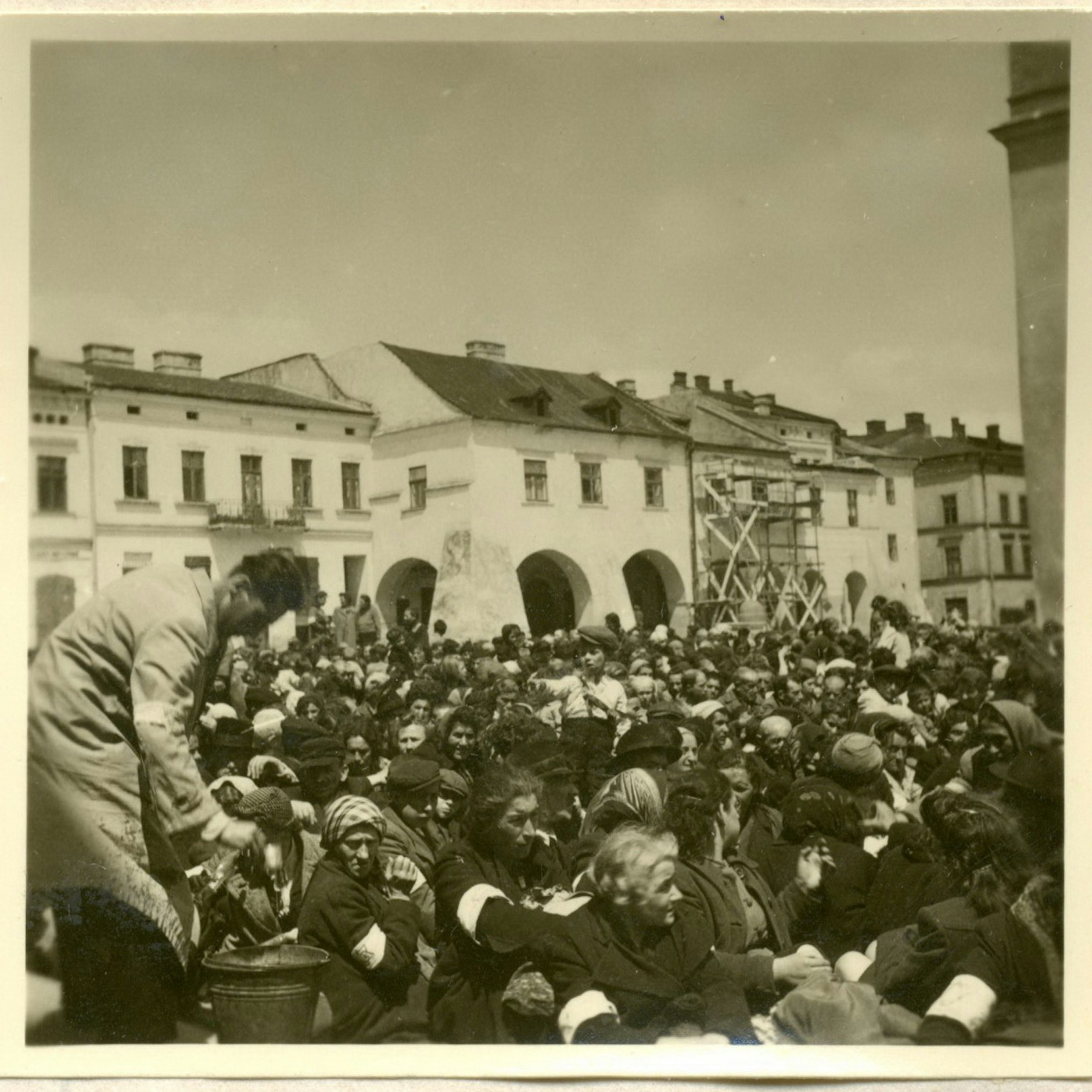 Die Festgehaltenen leiden Durst. Ein Mann mit Armbinde, der möglicherweise zum jüdischen Ordnungsdienst gehört, schöpft Wasser mit einem Becher aus einem herbeigebrachten Eimer.