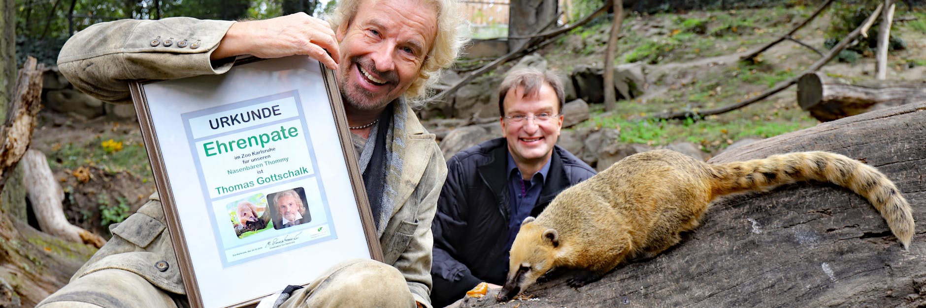 Thomas Gottschalk sitzt bei seinem Besuch im Zoo Karlsruhe mit Nasenbär „Thommy“ auf einem Baumstamm. Mit Abstand dahinter hockt Zoodirektor Matthias Reinschmidt.