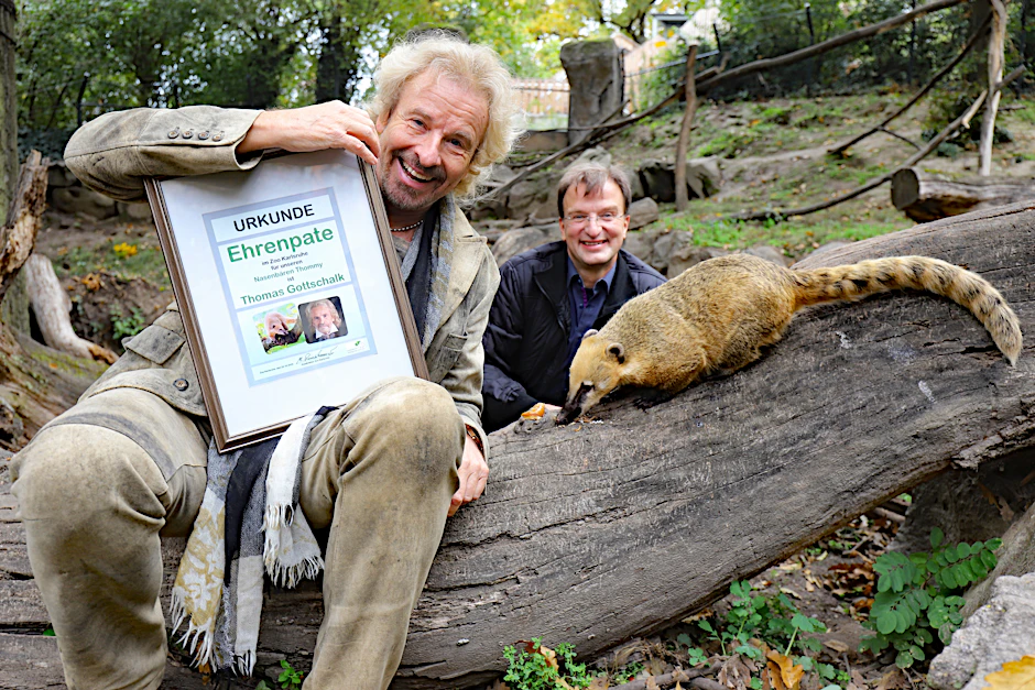 Thomas Gottschalk sitzt bei seinem Besuch im Zoo Karlsruhe mit Nasenbär „Thommy“ auf einem Baumstamm. Mit Abstand dahinter hockt Zoodirektor Matthias Reinschmidt.
