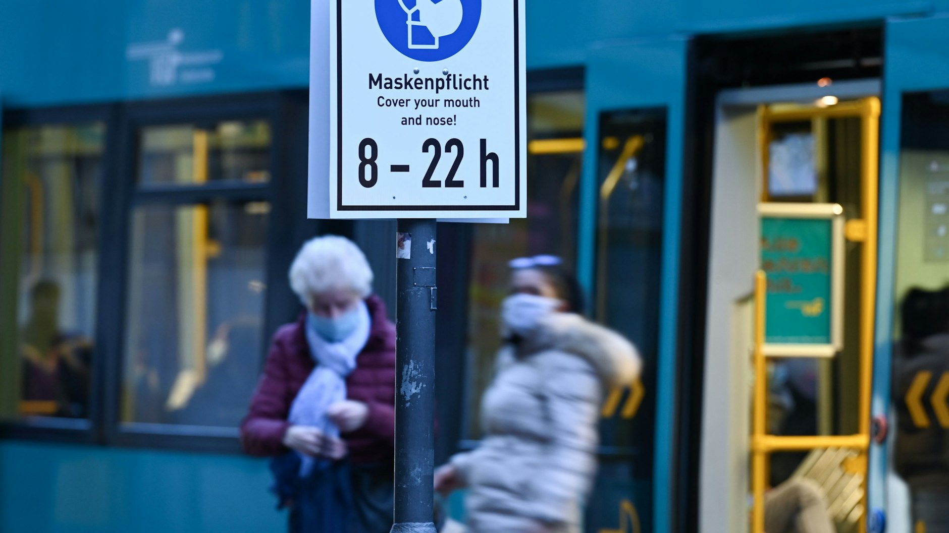 Fahrgäste mit Mund-Nasen-Schutz stehen am frühen Morgen vor einer haltenden Straßenbahn an einer Straßenbahnhaltestelle.