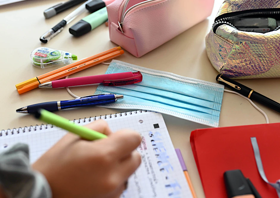 Eine Mund-Nase-Schutzmaske liegt zwischen Schreibutensilien auf einem Tisch, während ein Schüler einen Stift über einem Schreibblock hält.&nbsp;