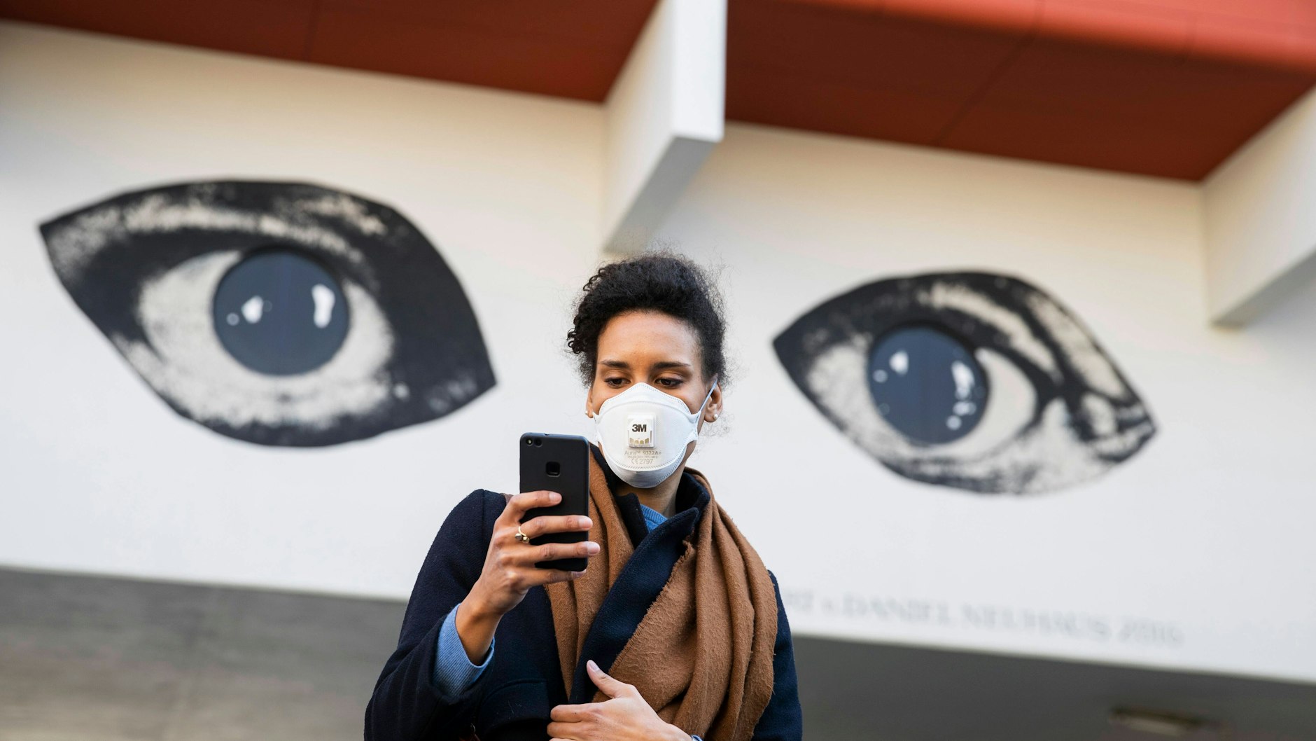 Eine junge Frau steht in der Schöneberger Goebenstraße und schaut auf ihr Handy mit den Augen des Grafittis am Pallasseum&nbsp; im Hintergrund.&nbsp;