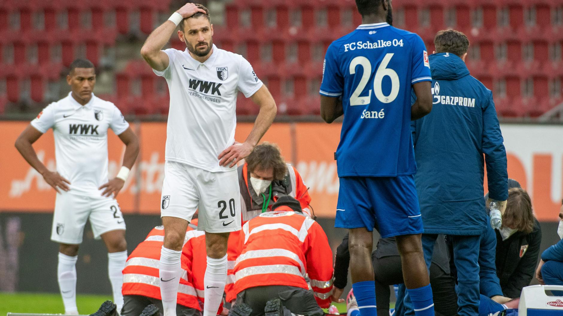 Augsburgs Daniel Caligiuri fasst sich an den Kopf, während Schalkes Mark Uth behandelt wird.