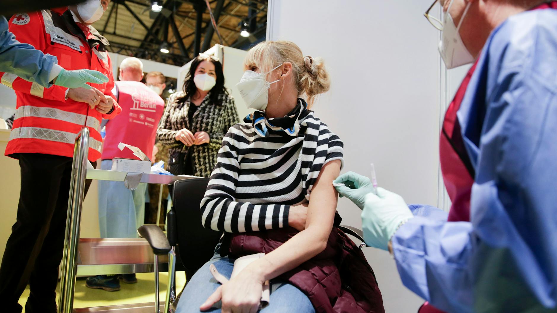Eine Frau, die in einem Pflegeheim arbeitet, wird im Impfzentrum Arena Treptow gegen das Coronavirus geimpft. Im Hintergrund steht Gesundheitssenatorin Dilek Kalayci.