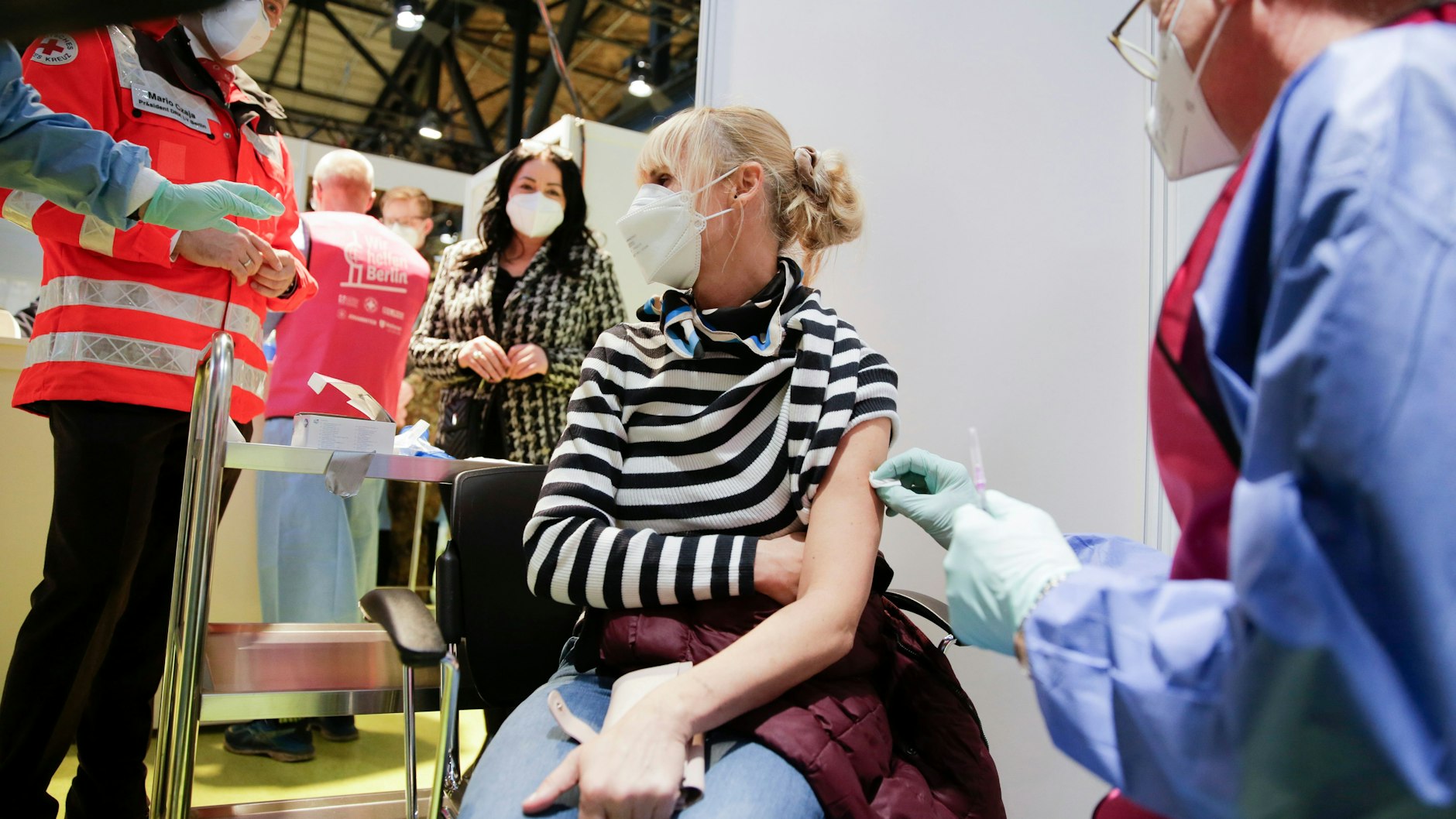 Eine Frau, die in einem Pflegeheim arbeitet, wird im Impfzentrum Arena Treptow gegen das Coronavirus geimpft. Im Hintergrund steht Gesundheitssenatorin Dilek Kalayci.