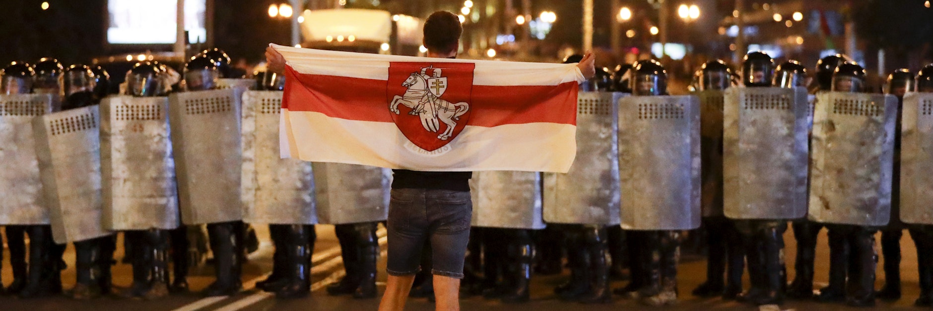 Ein Mann steht vor einer Wand von Polizeischilden. Er hält die Flagge Weißrusslands, die Lukaschenka 1995 abgeschafft hatte. Sie ist jetzt ein Symbol der Opposition.
