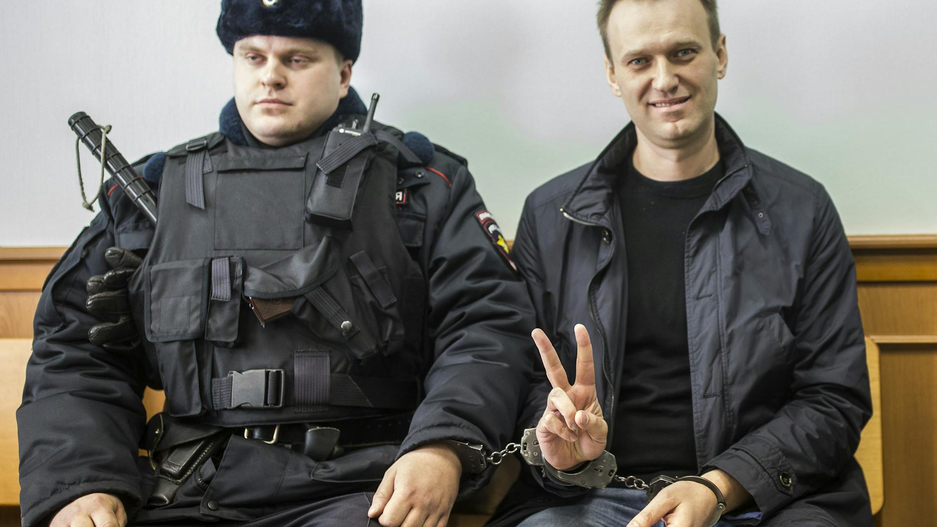 2017 musste Alexej Nawalny vor Gericht, zeigt ein Victory-Zeichen, während er neben einem Sicherheitsbeamten sitzt. 