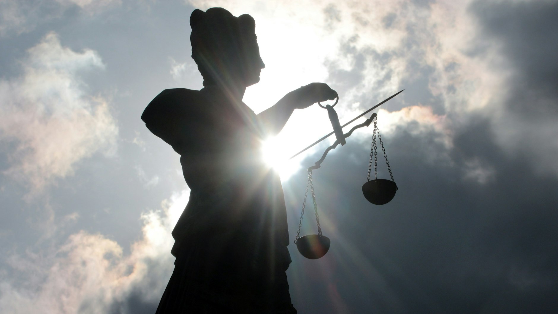 Silhouette der Justitia Skulptur mit Schwert und Waagschalen vor schwarzen, düsteren Wolken und Gegenlicht.