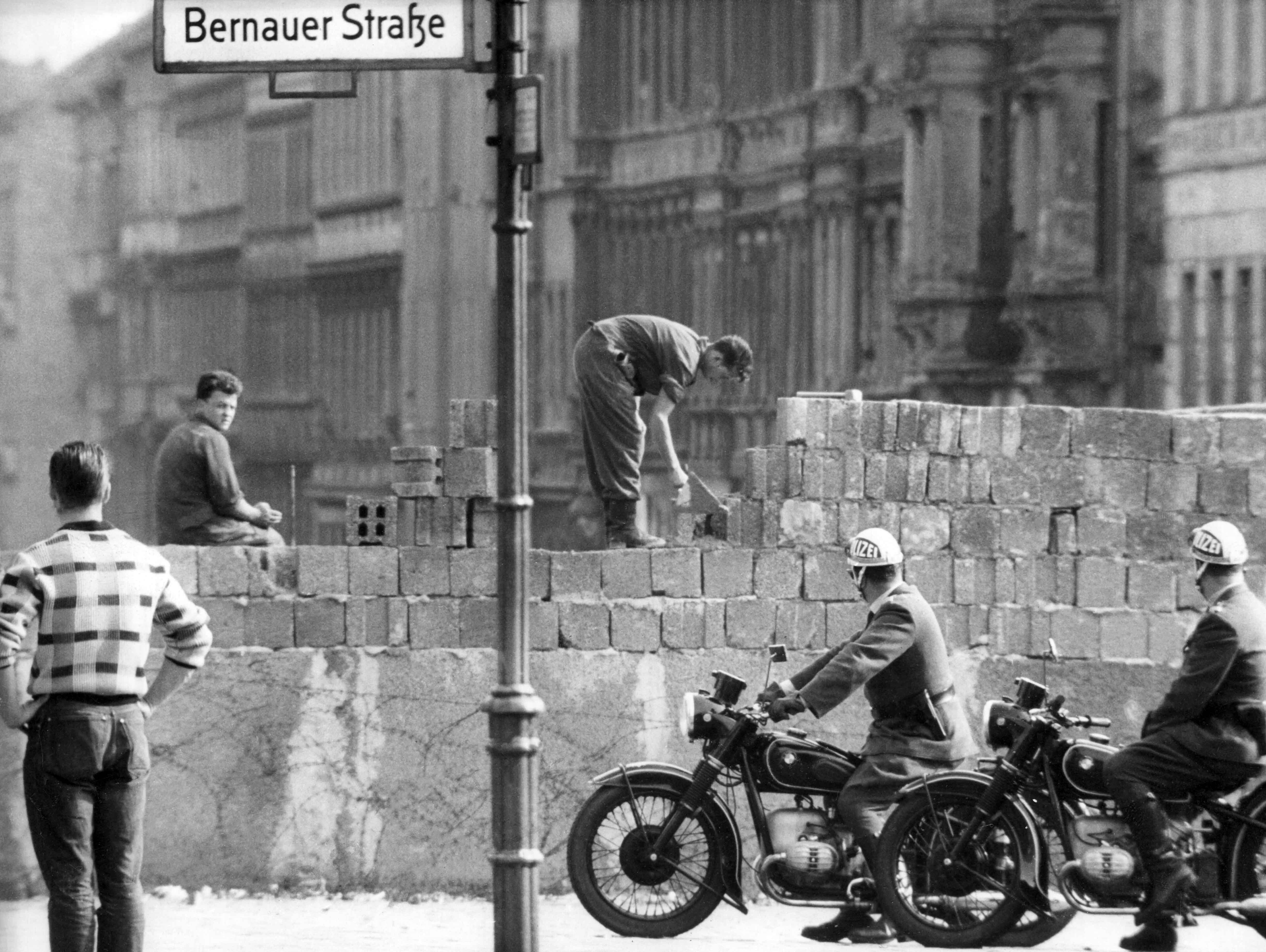 Mauerbau 1961: Stein für Stein wächst die Mauer, die Ost-Berliner Arbeiter im August des Jahres an der Bernauer Straße errichten. West-Berliner Polizisten und Passanten sehen dem ungläubig zu.
