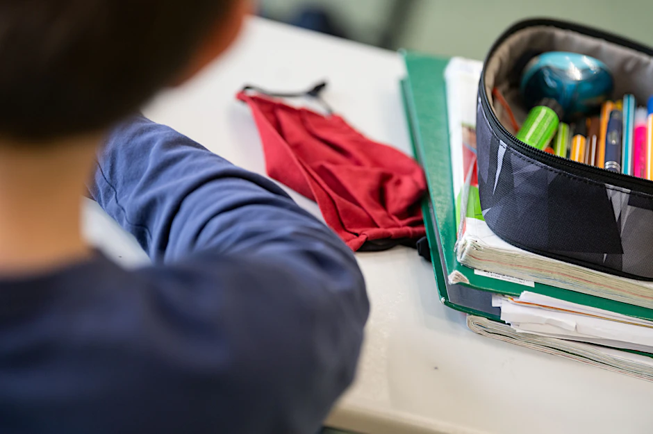 Eine Mund-Nasen-Bedeckung liegt während einer Unterrichtsstunde neben einem Mäppchen und Schulbüchern auf einem Schultisch.