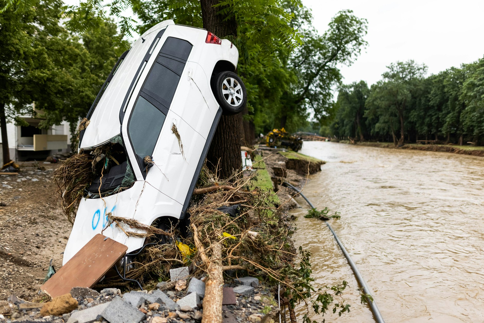Ein vom Hochwasser angeschwemmtes Auto lehnt an einem Baum in Bad Neuenahr während im Hintergrund der Fluss Ahr zu sehen ist.