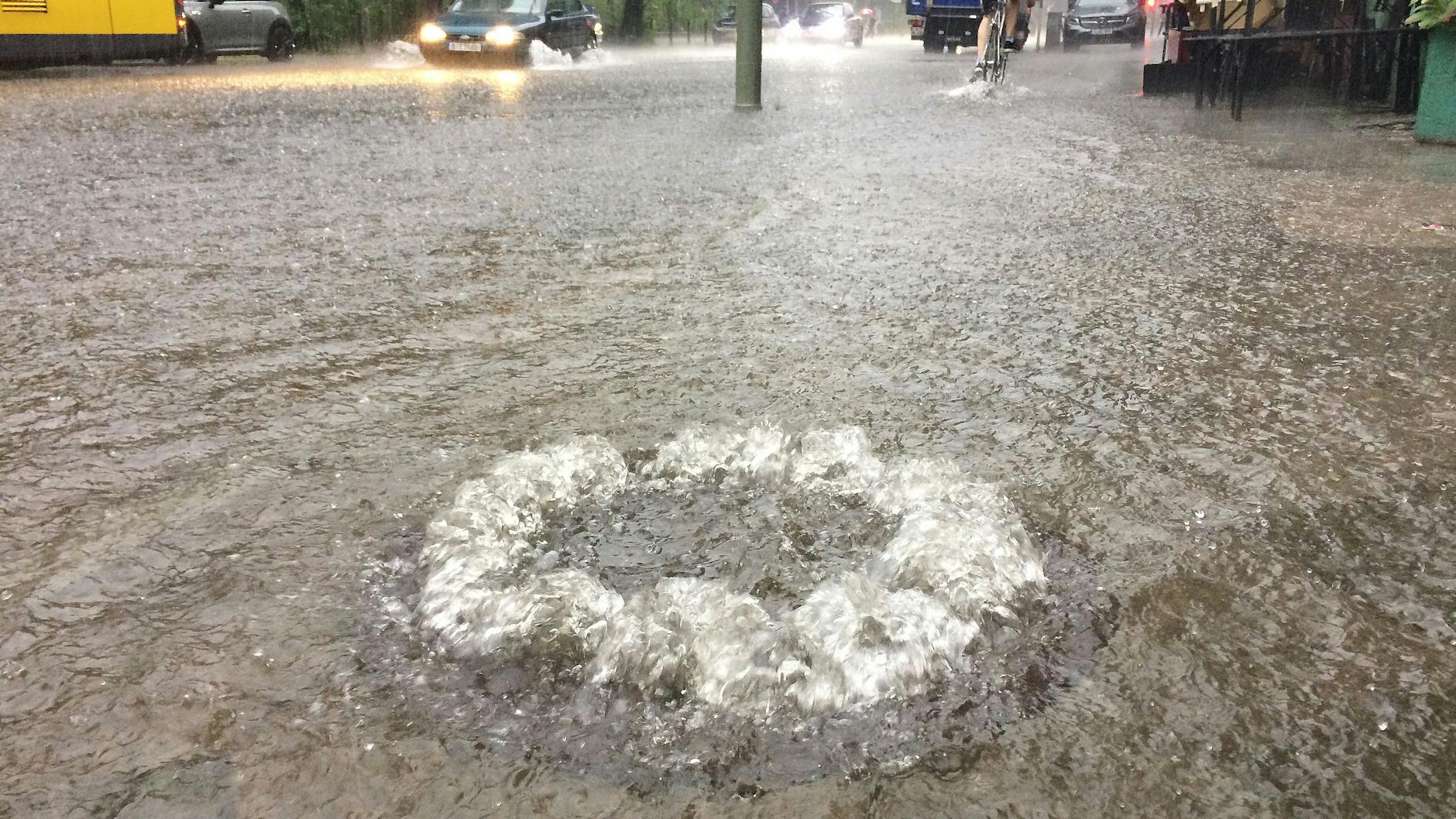 Nach Starkregen sprudelt Wasser auf einer überfluteten Straße im Stadtteil Schöneberg aus einem Gully.