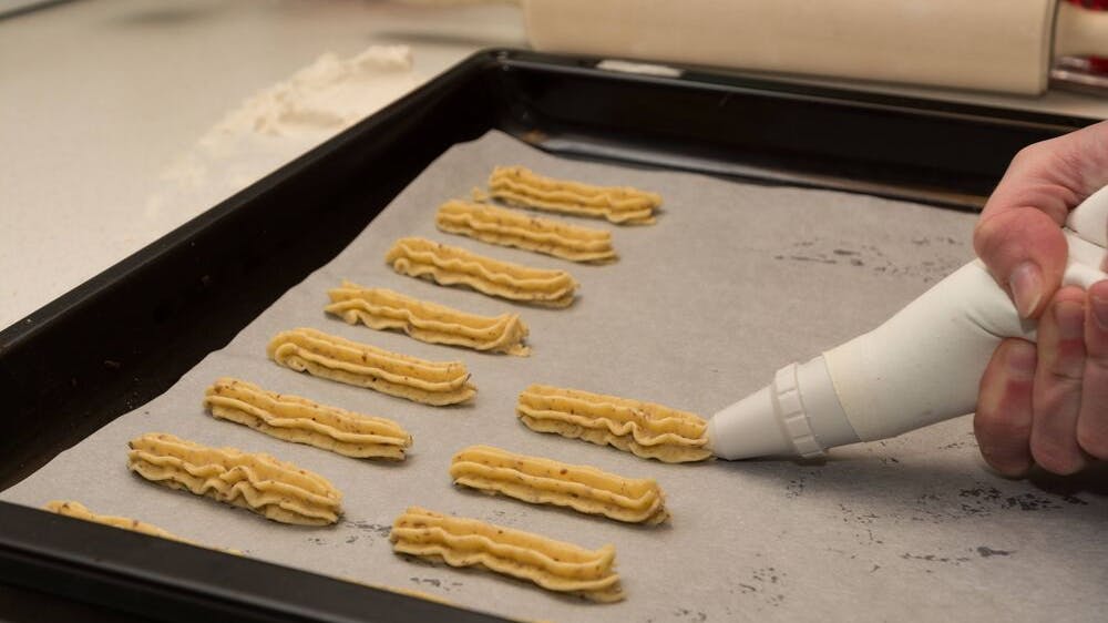 Mit einem Spritzbeutel wird der Teig für das Spritzgebäck auf dem Backblech in ca. 5 Zentimeter langen Streifen gespritzt und bekommt so seine typische Form.