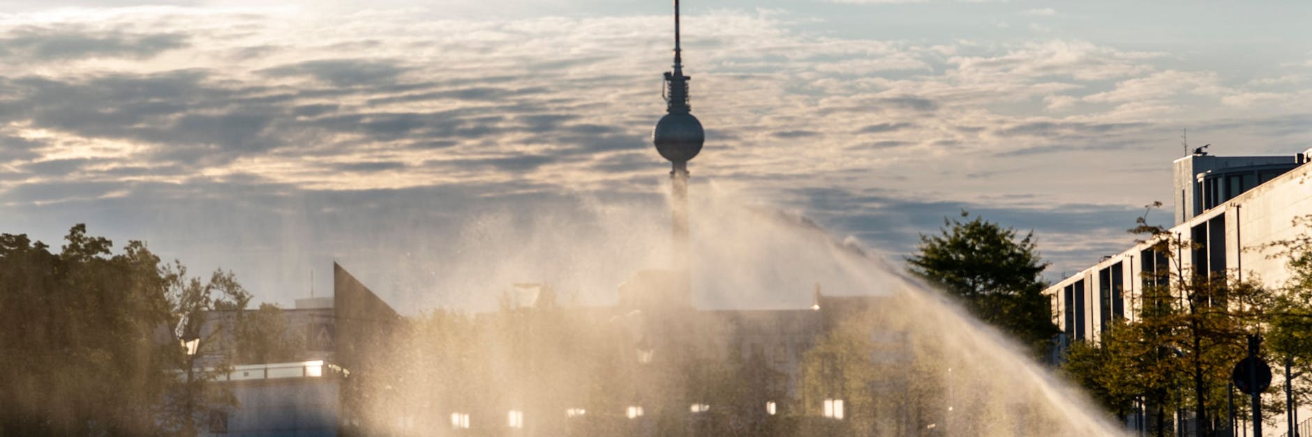 Es wird wieder glutheiß: Das Wasser eines Rasensprengers berieselt im Gegenlicht der Sonne im Regierungsviertel eine Rasenfläche und Bäume.