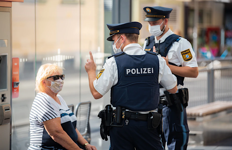 Zwei Polizisten nehmen an einer Tram-Haltestelle in Bayern&nbsp;die Personalien einer Bürgerin auf.&nbsp;Die Frau soll gegen die Maskenpflicht im öffentlichen Nahverkehr verstoßen haben.