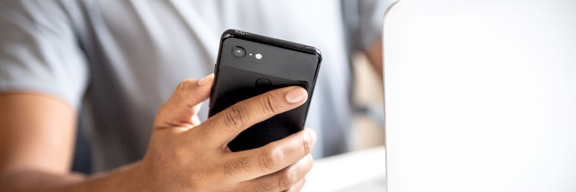 Ein junger Mann sitzt an einem Schreibtisch am Laptop und checkt sein Smartphone.