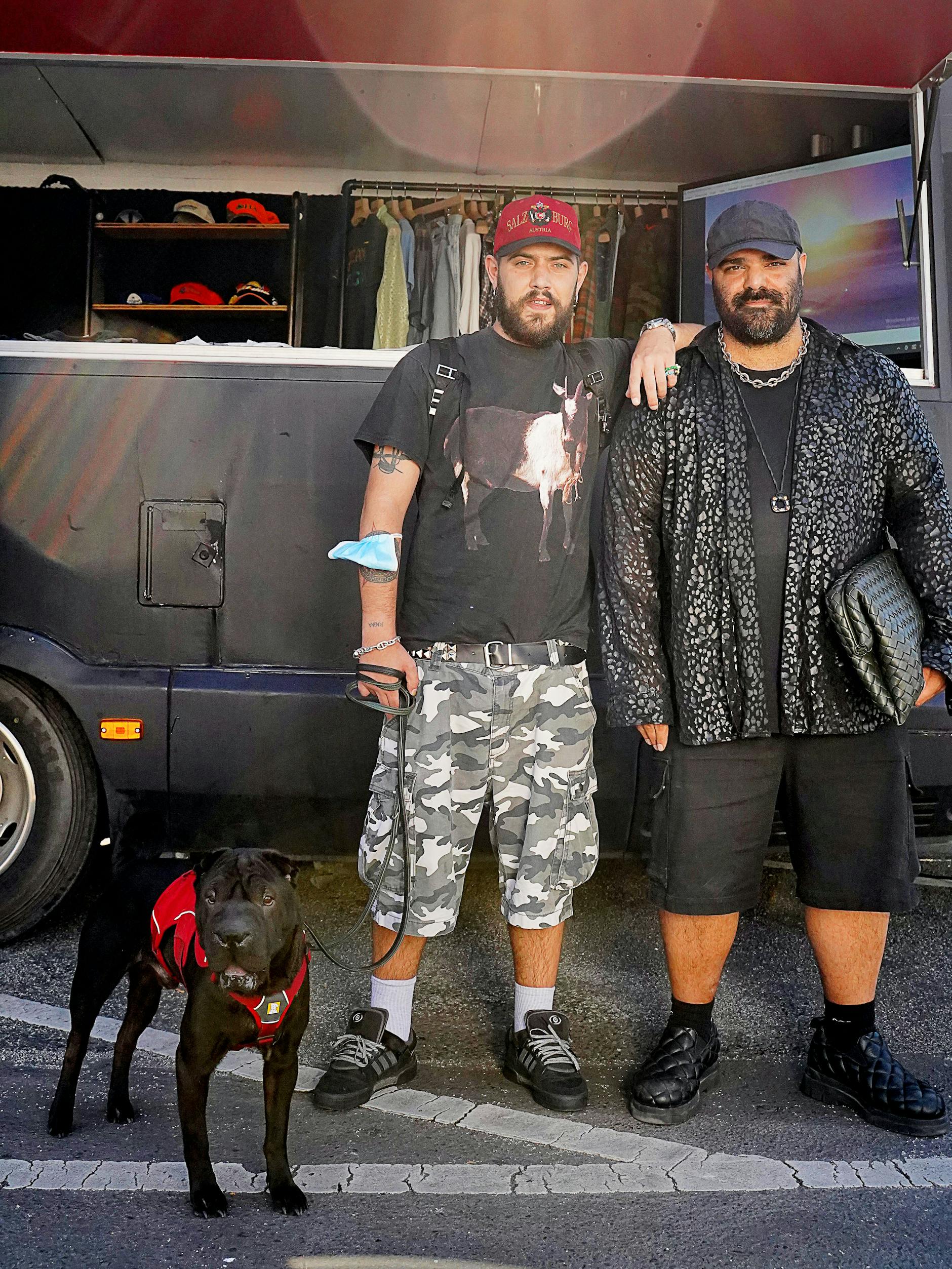 Mit Heiligenschein: Das Power-Couple Marc und Mumi samt Hund Ragna. Marc arbeitet als Fashion Director bei 032c, Mumi ist der Veranstalter des Reference Festivals und CEO der gleichnamigen Agentur. Marcs Outfit: Souvenir-Basecap aus Salzburg, Merchandise-Shirt von Adam Sandler, Carhartt-Shorts und adidas x Bad Bunny. Mumis Look: Cap von Our Legacy, Gildan-T-Shirt, Hemd von Black Comme des Garçons, Socken von 032c sowie Shorts und Schuhe von Bottega Veneta. Dazu Ketten von Marni und Nhat-Vu Dang.