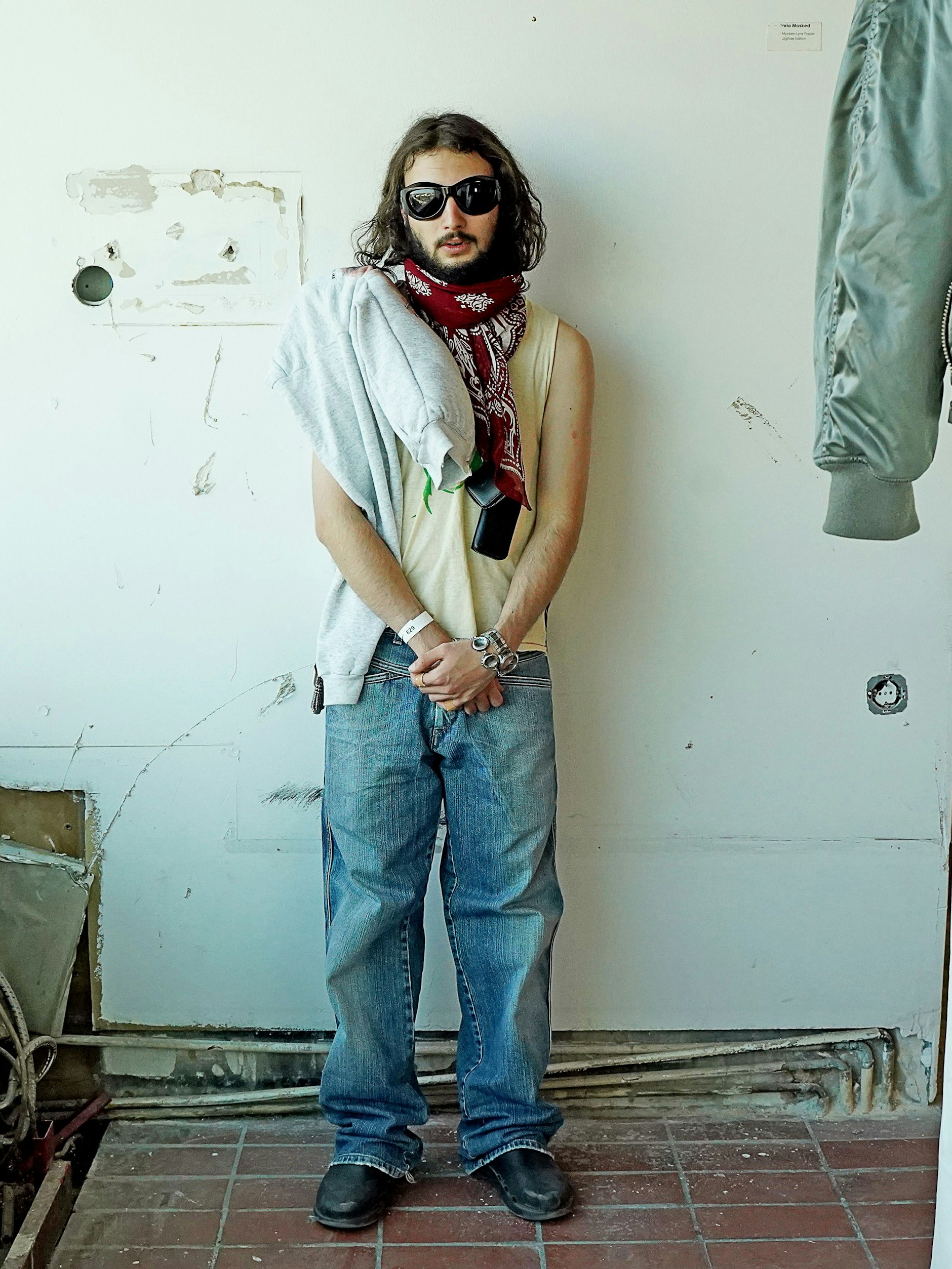 Deniz neben seiner Installation im Bierpinsel. Der Künstler und Mitbegründer des Berliner Labels Treffpunkt trägt ein Tanktop vom japanischen Label Undercover, eine Jeans von Marithé+François Girbaud und Sneakers von Oofos. Das Sweatshirt über seiner Schulter ist von Comme des Garçons, die Brille von Bottega Veneta.