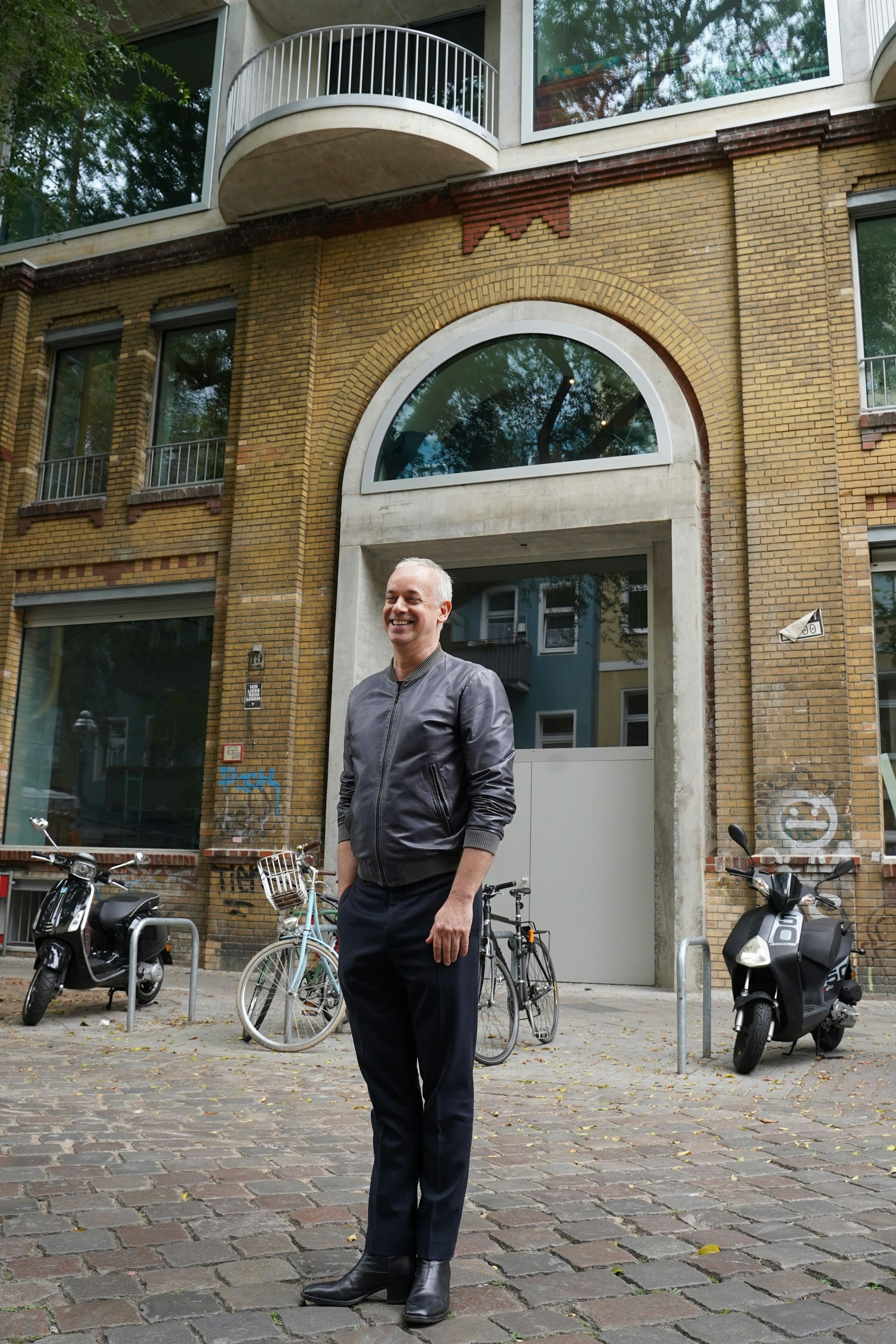 So gut gelaunt kann nur jemand sein, der es geschafft hat! Der Architekt Thomas vor seinem Werk, <em>dem</em> Hingucker in der Malplaquetstraße. Durch die Einbindung der alten Einfahrt zu den ehemaligen Osram-Höfen in den Neubau schmiegt sich das Wohnhaus perfekt zwischen die übrigen Fassaden und verleiht der Straße gleichzeitig einen zeitgenössischen Touch. Thomas trägt Prada (Jacke) und Jil Sander (Hose).