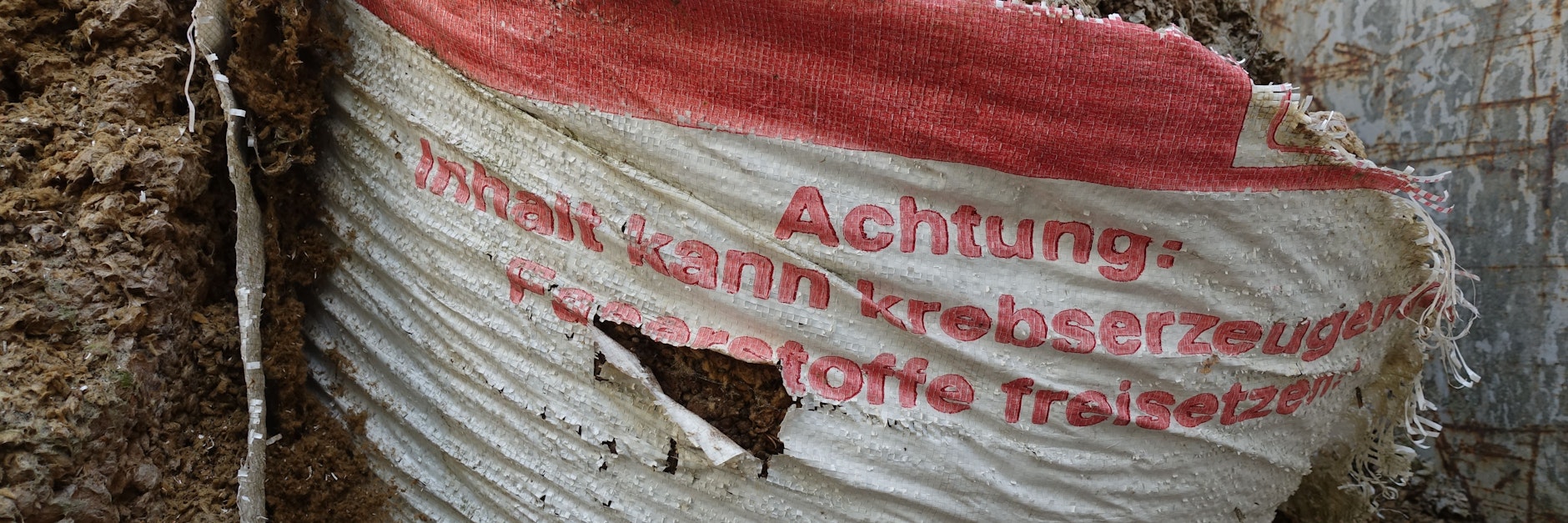 Auf dem Gelände liegen in Schuttcontainern aufgerissene Säcke mit der Aufschrift: „Achtung: Inhalt kann krebserzeugende Faserstoffe freisetzen“.