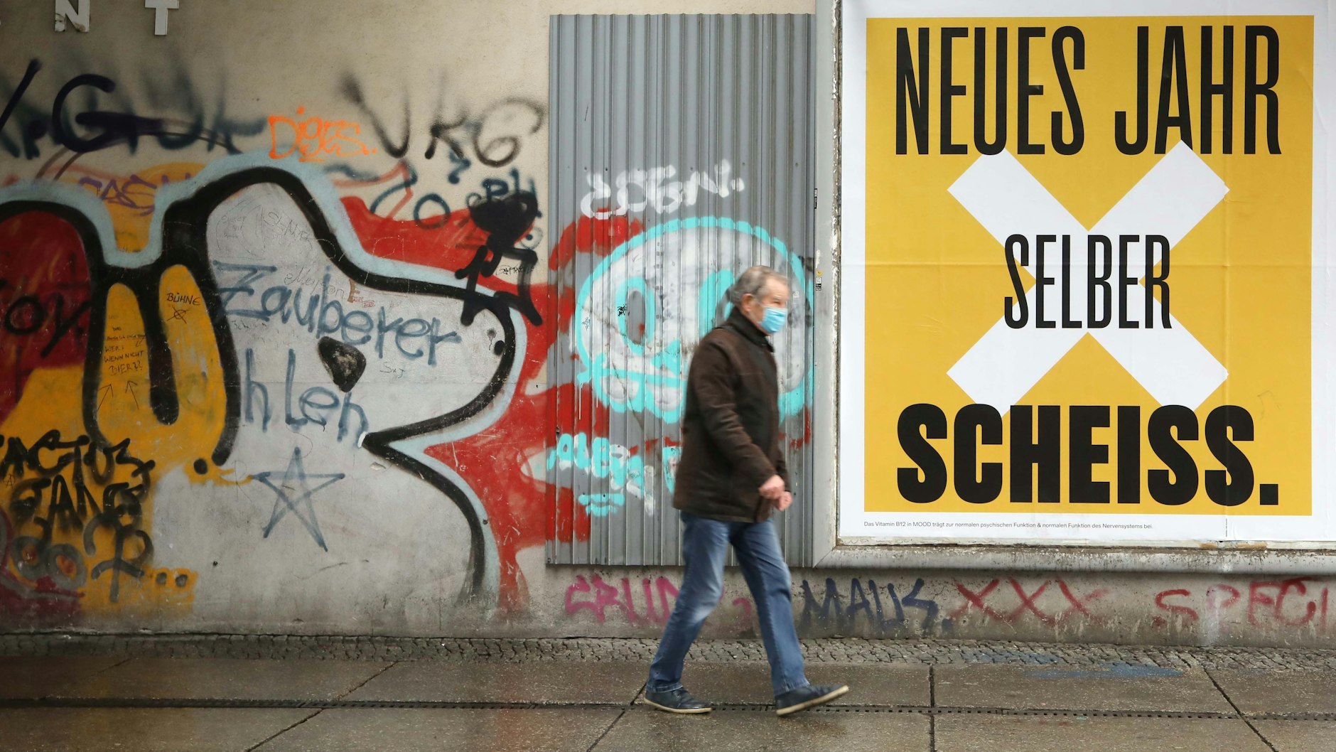 Ein Mann mit Maske spaziert in Berlin an einer beklebten Hauswand vorbei.&nbsp;
