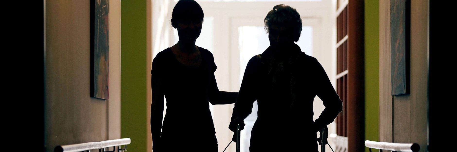 Eine Pflegekraft begleitet die Bewohnerin eines Altenheims mit Rollator beim Gang durch den Flur. 