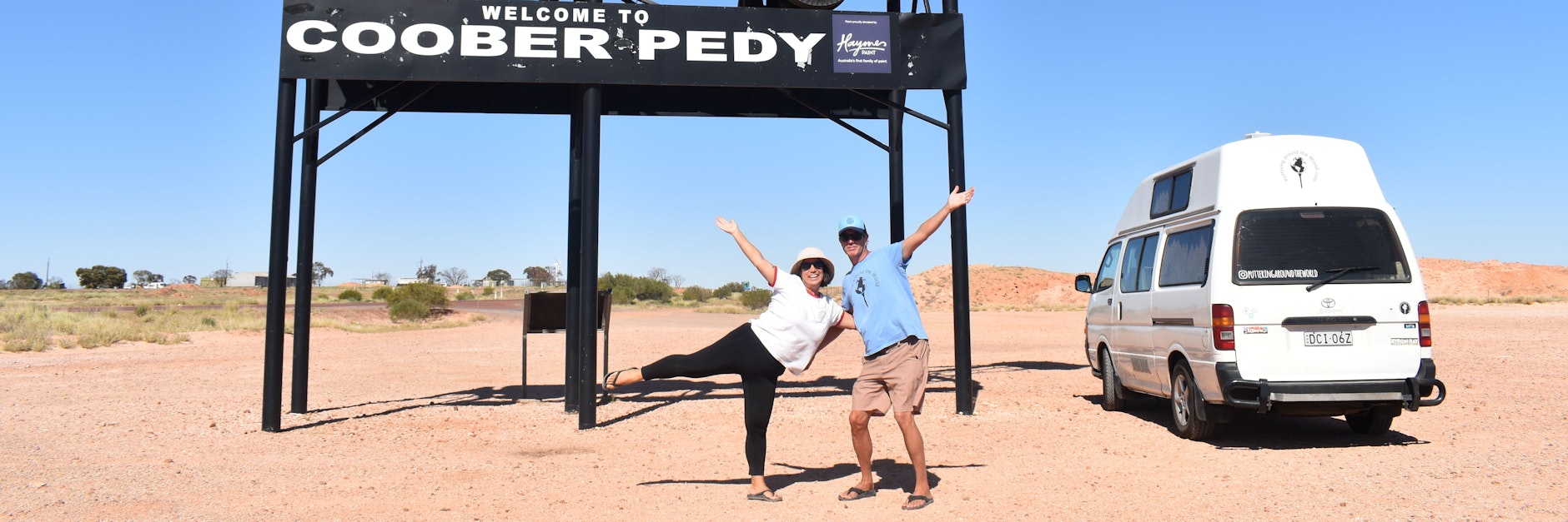 Menekse (li.) und James Humphrey stehen vor einem Ortsschild im australischen Coober Pedy neben ihrem ausgebauten Van, den sie liebevoll „Putu“ nennen. Seit zwei Jahren ist er ihr Fahrzeug und ihr Zuhause.