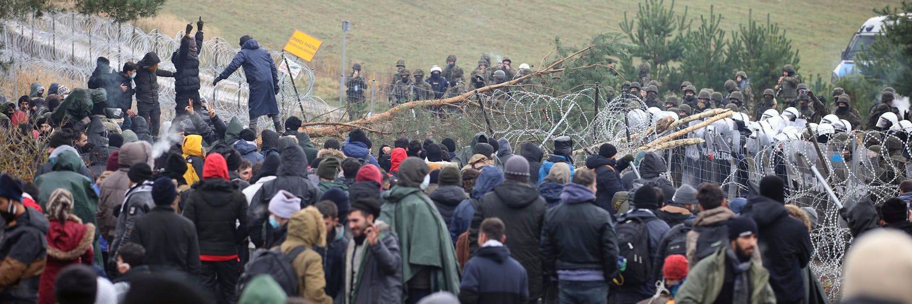  Polnische Polizisten stehen hinter einem Stacheldrahtzaun, während sich an der belarussisch-polnischen Grenze Migranten versammelt haben. 