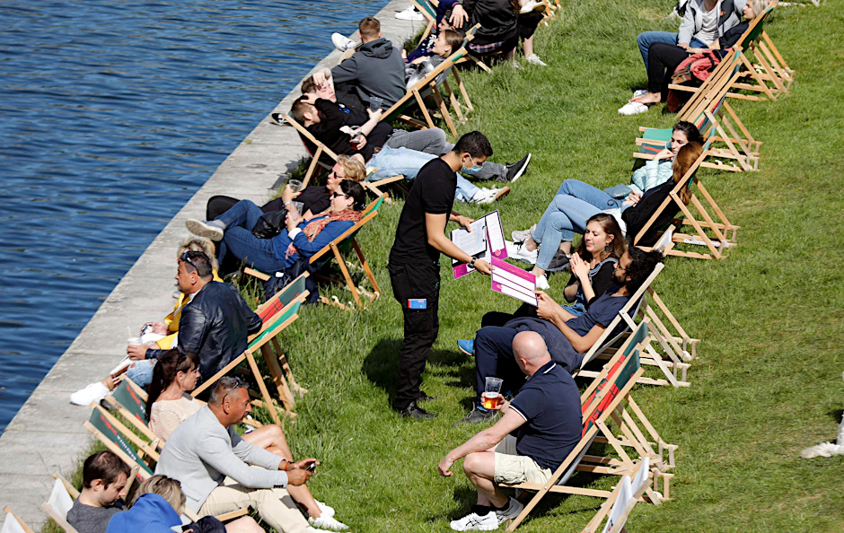 Ein Ober mit Gesichtsmaske bedient Gäste in einem gut besuchten Café an der Spree. So sah der Weg aus dem ersten Lockdown im Mai vorigen Jahres aus. Und so könnte es bald wieder kommen.