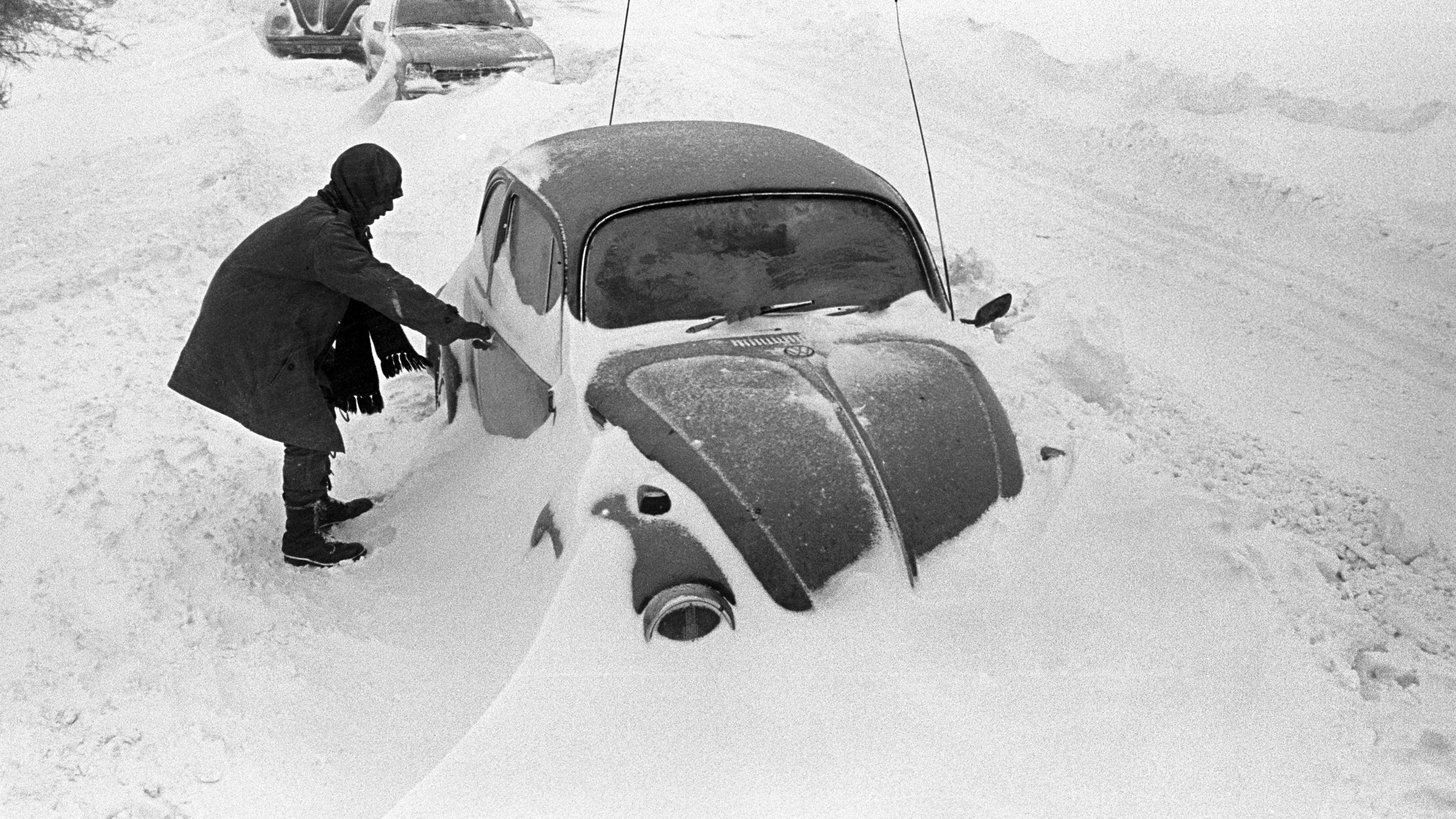 Ein Autofahrer steht in Cuxhaven (Niedersachsen) neben seinem am Straßenrand im Schnee steckengebliebenen VW-Käfer.