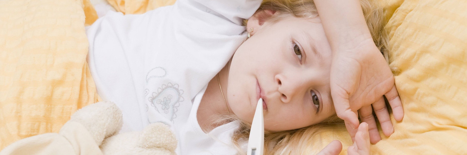 Ein Mädchen liegt mit Fieberthermometer im Mund im Bett.