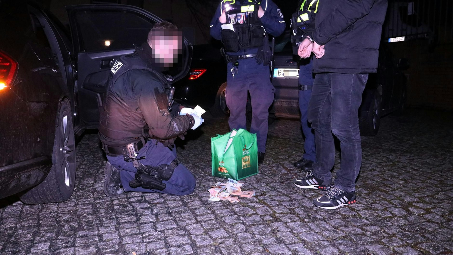 Ein Polizist zählt das gefundene Bargeld, während einer der Verdächtigen danebensteht.&nbsp;
