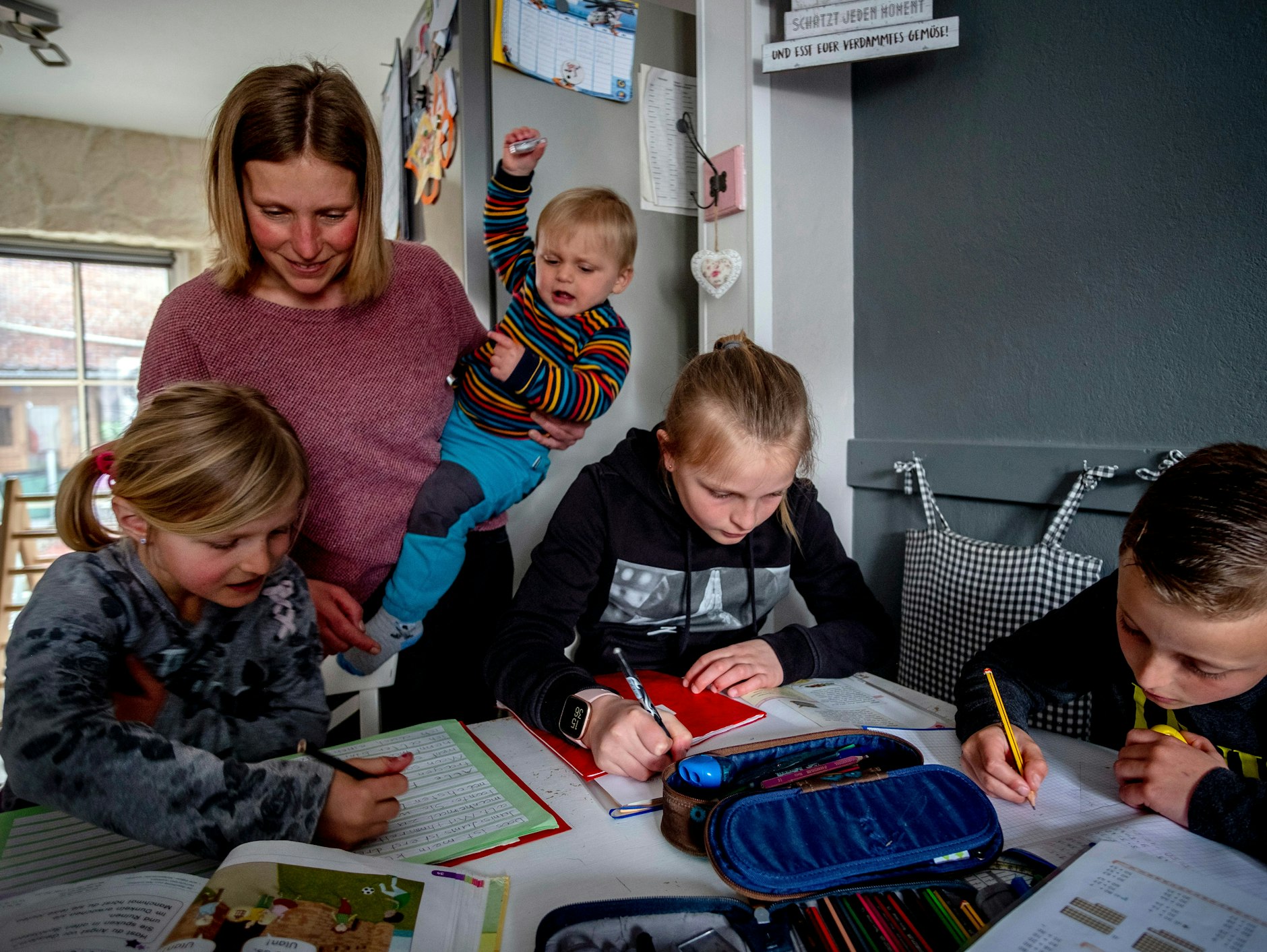Eine Mutter, das ungebärdige Kleinkind auf dem Arm, hilft ihren Schulkindern bei den Aufgaben. Vielleicht läuft nebenan eine Video-Konferenz, die sie gerade versäumt. 