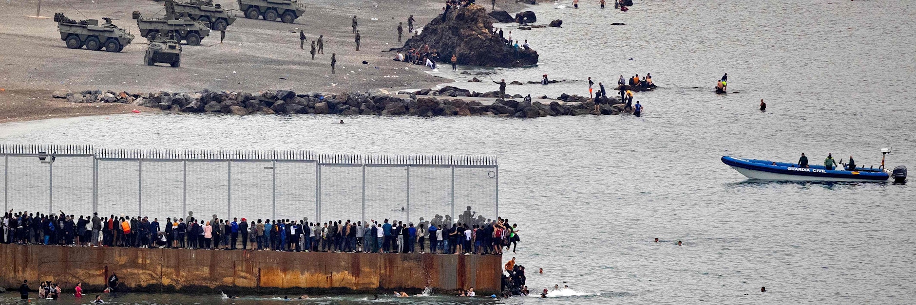 Menschen drängen sich auf einer ins Meer gebauten Mole mit dem Grenzzaun, springen ins Wasser, um das spanische Ceuta zu erreichen. Dort sind Radpanzer aufgefahren.