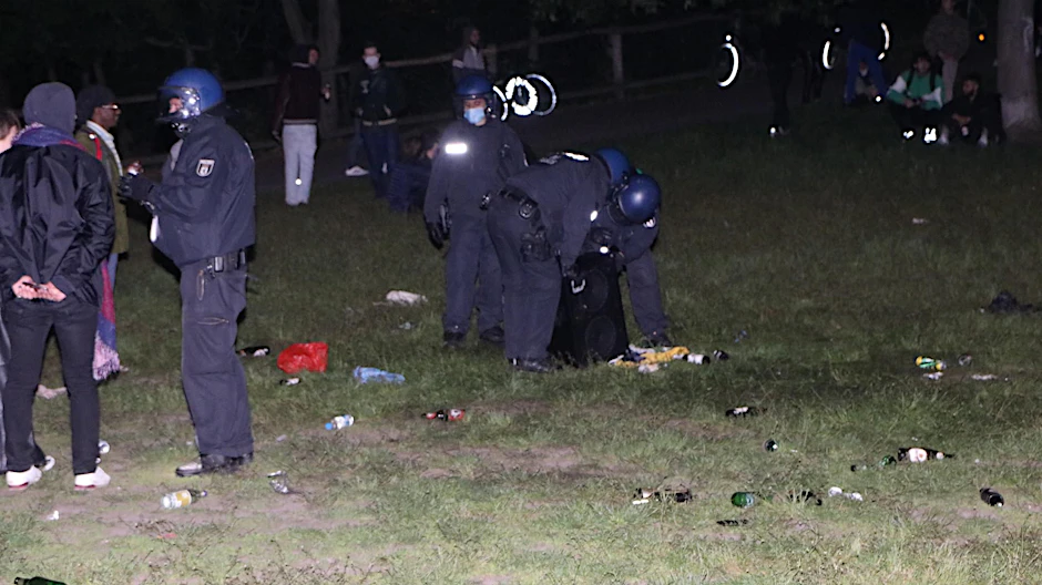 Polizisten heben in der Hasenheide eine Musikanlage hoch, umgeben von Flaschen und anderem Müll.