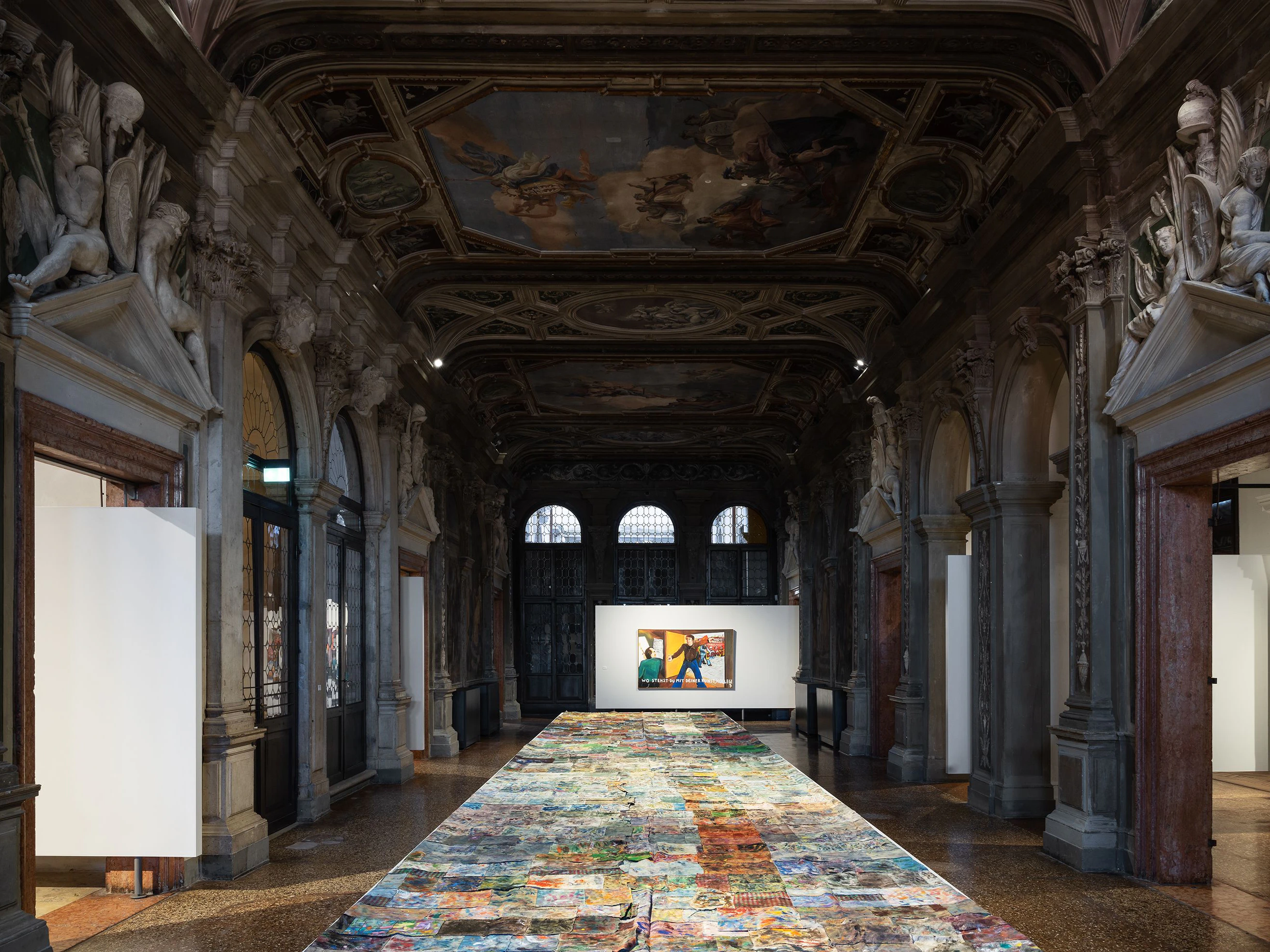In Pradas Venedig-Dependance läuft die Ausstellung „Stop Painting“. Auf dem Boden ein Werk von  Jean-Frédéric Schnyder Hudel, hinten das Gemälde „Wo stehst du mit deiner Kunst, Kollege?“ von Jörg Immendorff, 1973.<br>