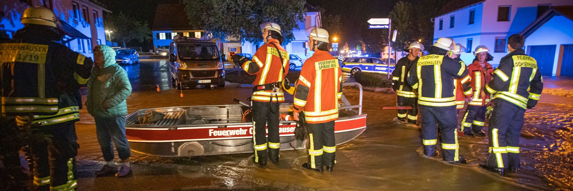 Feuerwehrleute stehen in Rot an der Rot neben einem Feuerwehrboot auf einer Straße in den Fluten eines heftigen Unwetters. Hier ertrank ein Mann in seiner Wohnung.