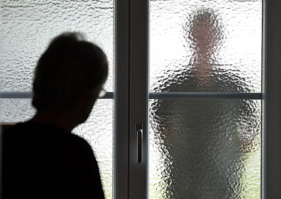 Eine Frau steht an einer Glasscheibe, hinter der ein Stalker wartet.&nbsp;