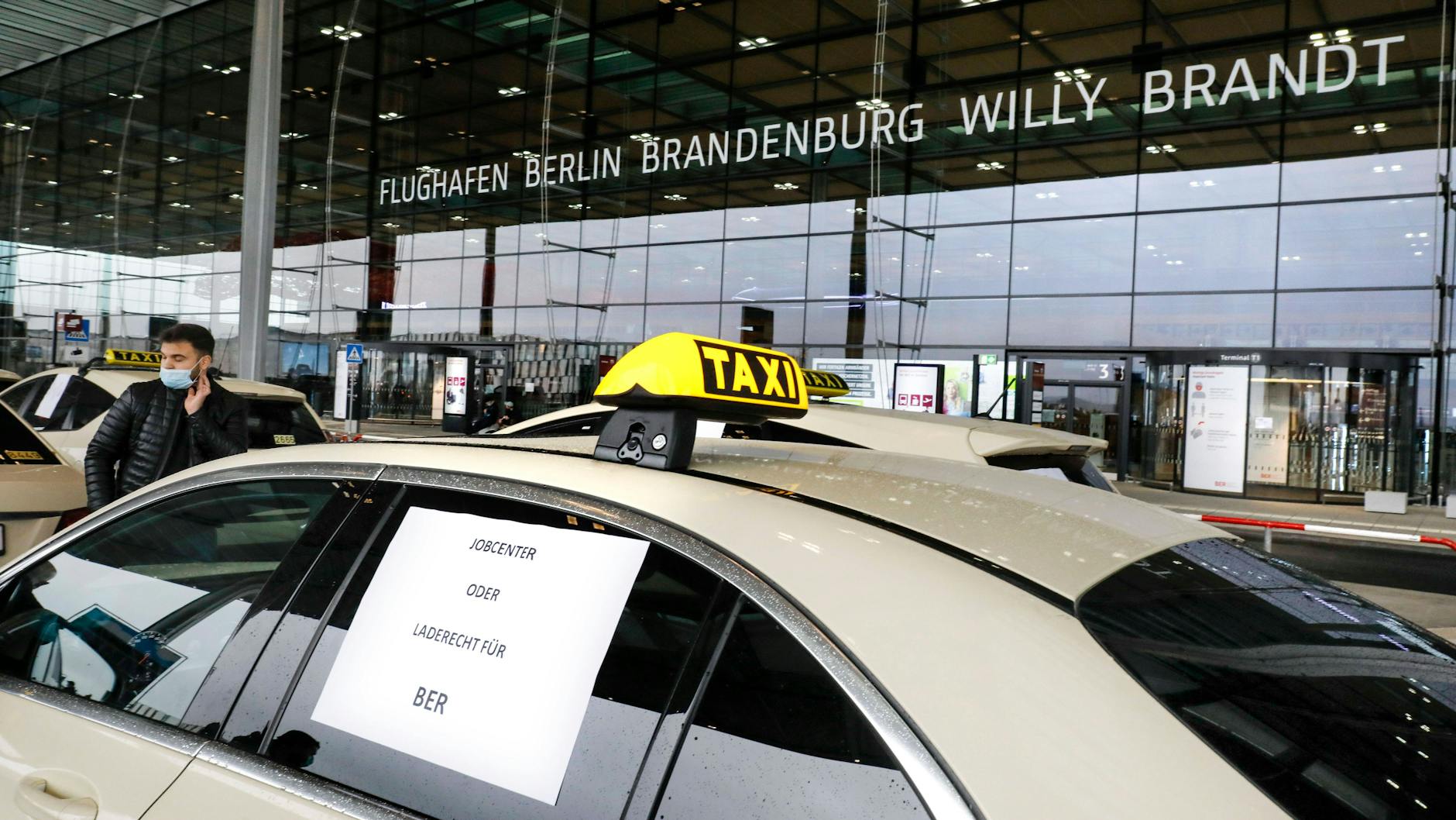 Ein Taxi am Flughafen BER. „Jobcenter oder Laderecht für BER“: Auf dem Blatt unterstreicht der Fahrer die Forderung, dass alle Berliner Taxis das Recht haben sollen, Fahrgäste am Flughafen aufzunehmen – nicht nur bis zu 400 wie derzeit.